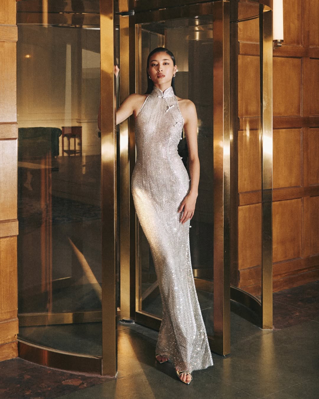 A floor-length, form-fitting halter-neck gown in silver sequins. It features a high mandarin collar with traditional Chinese-style frog closures on the chest.