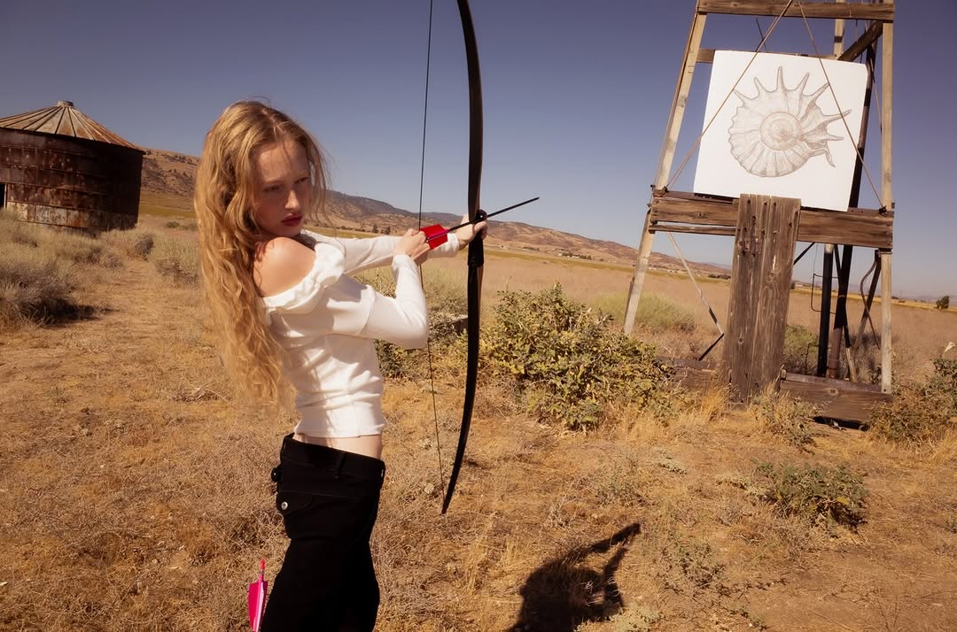 Model wearing a white long-sleeved off-the-shoulder crop top and black pants while holding a bow.