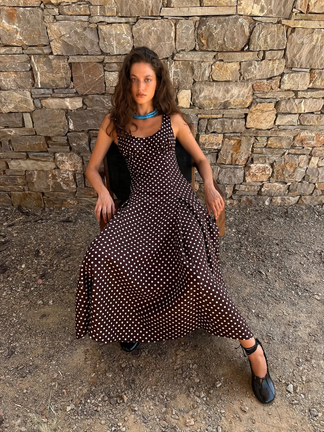 A model sits in a chair wearing a sleeveless brown midi dress with white polka dots.