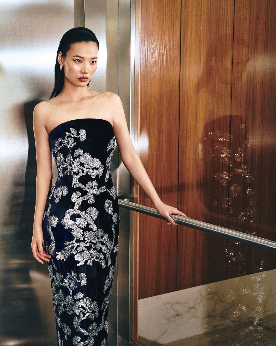 A model wearing a strapless midnight blue velvet gown featuring silver sequined pine tree embroidery.