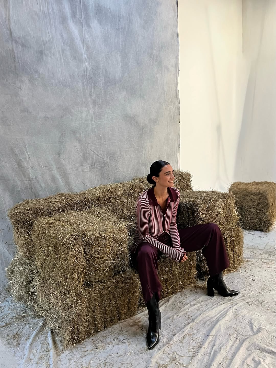 Full-body shot of model sitting on hay bales wearing a maroon and white striped shirt with solid collar and wide-leg maroon trousers.