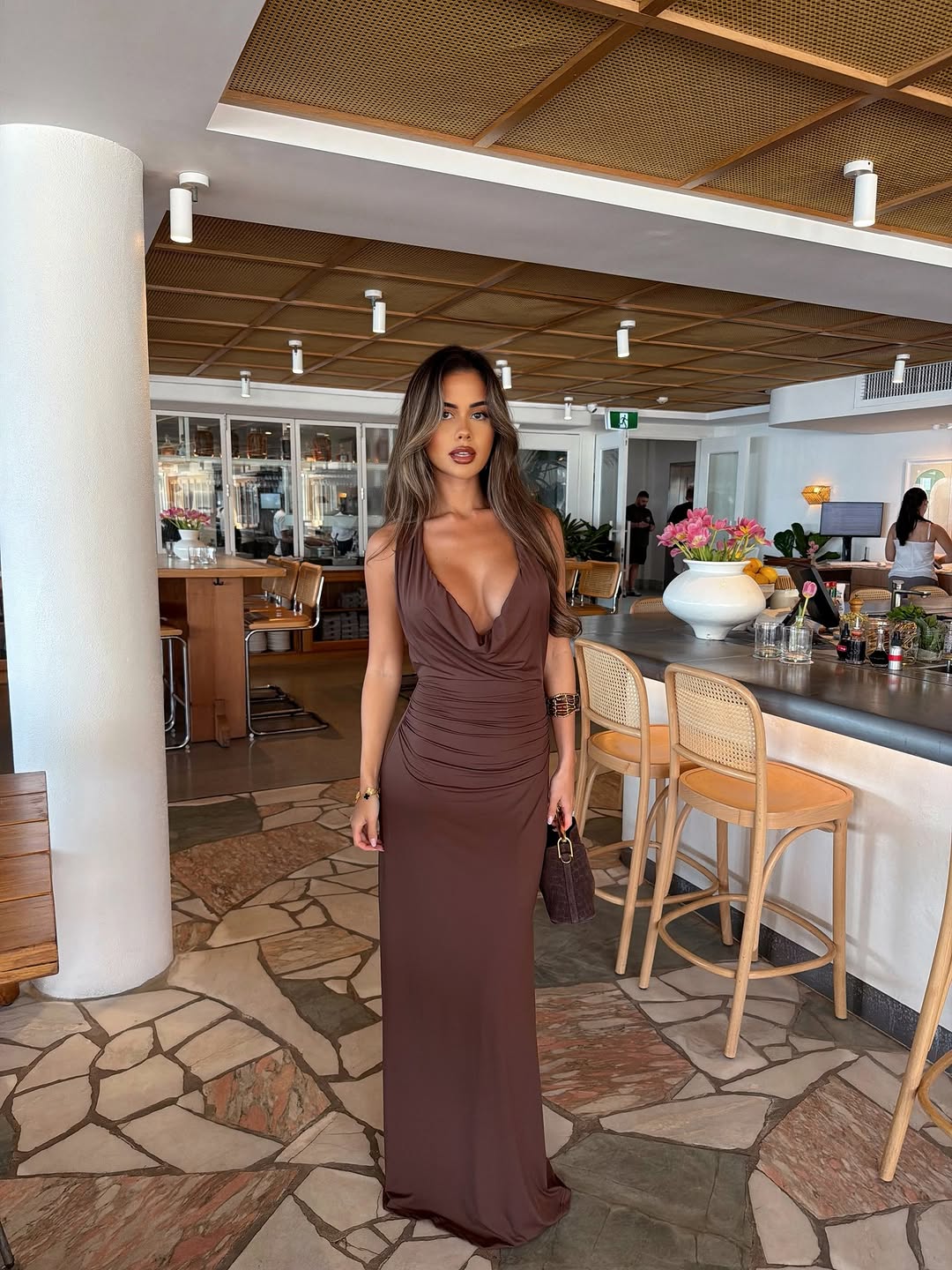 Front view of a woman in a long, brown sleeveless dress with a deep cowl neckline and ruched waist.