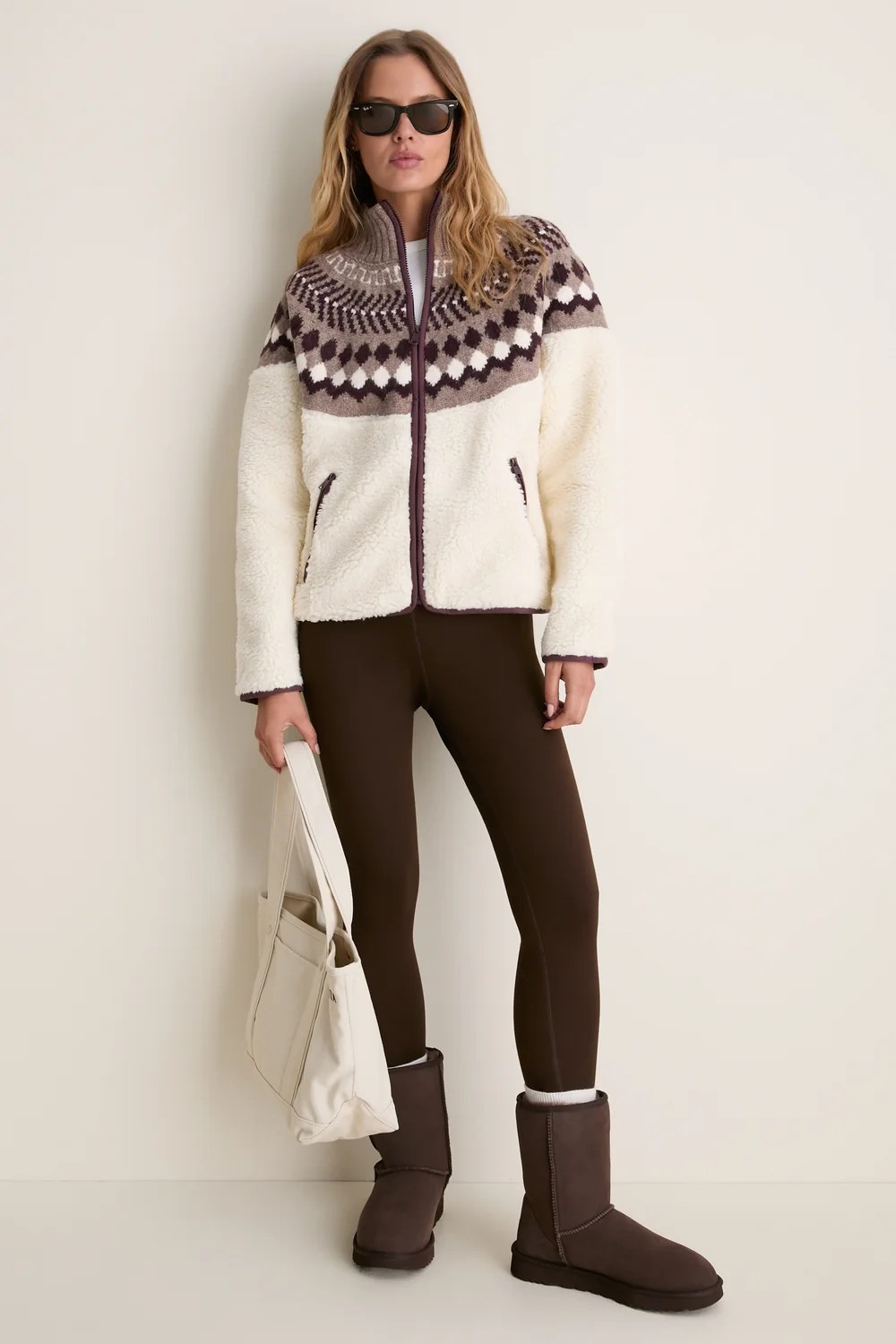 A cream-colored sherpa fleece zip-up jacket featuring a brown and white Fair Isle knit pattern across the shoulders, chest, and collar. It has dark brown trim along the zipper…
