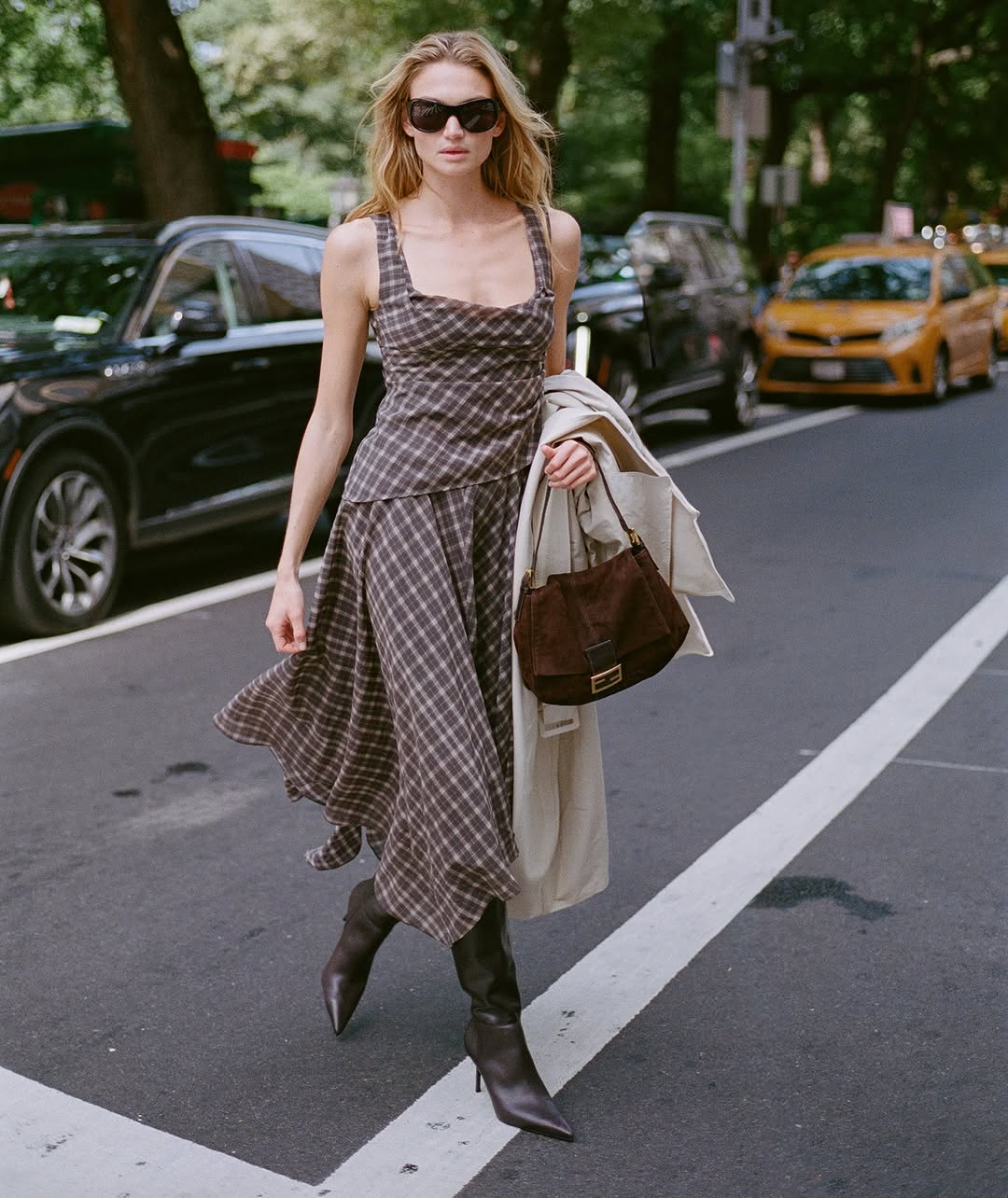 Full-body shot of a woman walking on a street wearing a brown plaid camisole top and matching midi skirt, paired with dark knee-high boots.