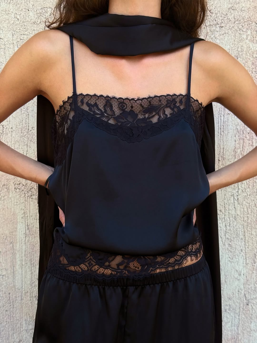Close-up of a black satin camisole with floral lace trim at the neckline and hem, worn with matching trousers.
