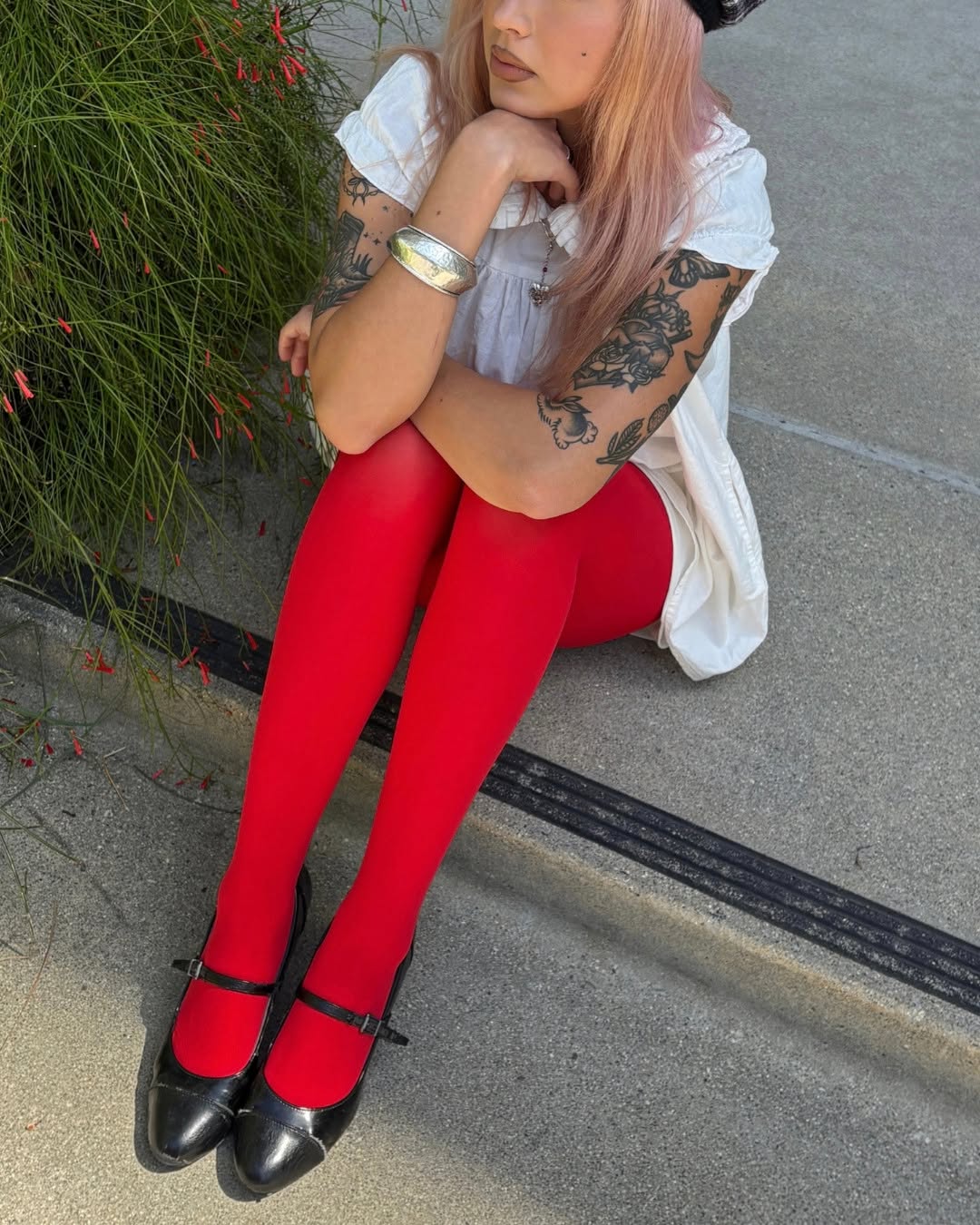 Model sitting on concrete wearing a white ruffled top, bright red tights, and black Mary Jane heels.