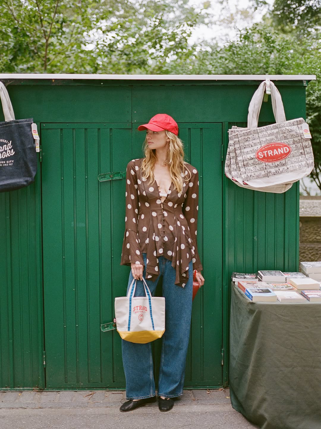 Full-body shot of a woman wearing a sheer brown long-sleeved blouse with white polka dots, blue wide-leg jeans, black shoes, and a red baseball cap.