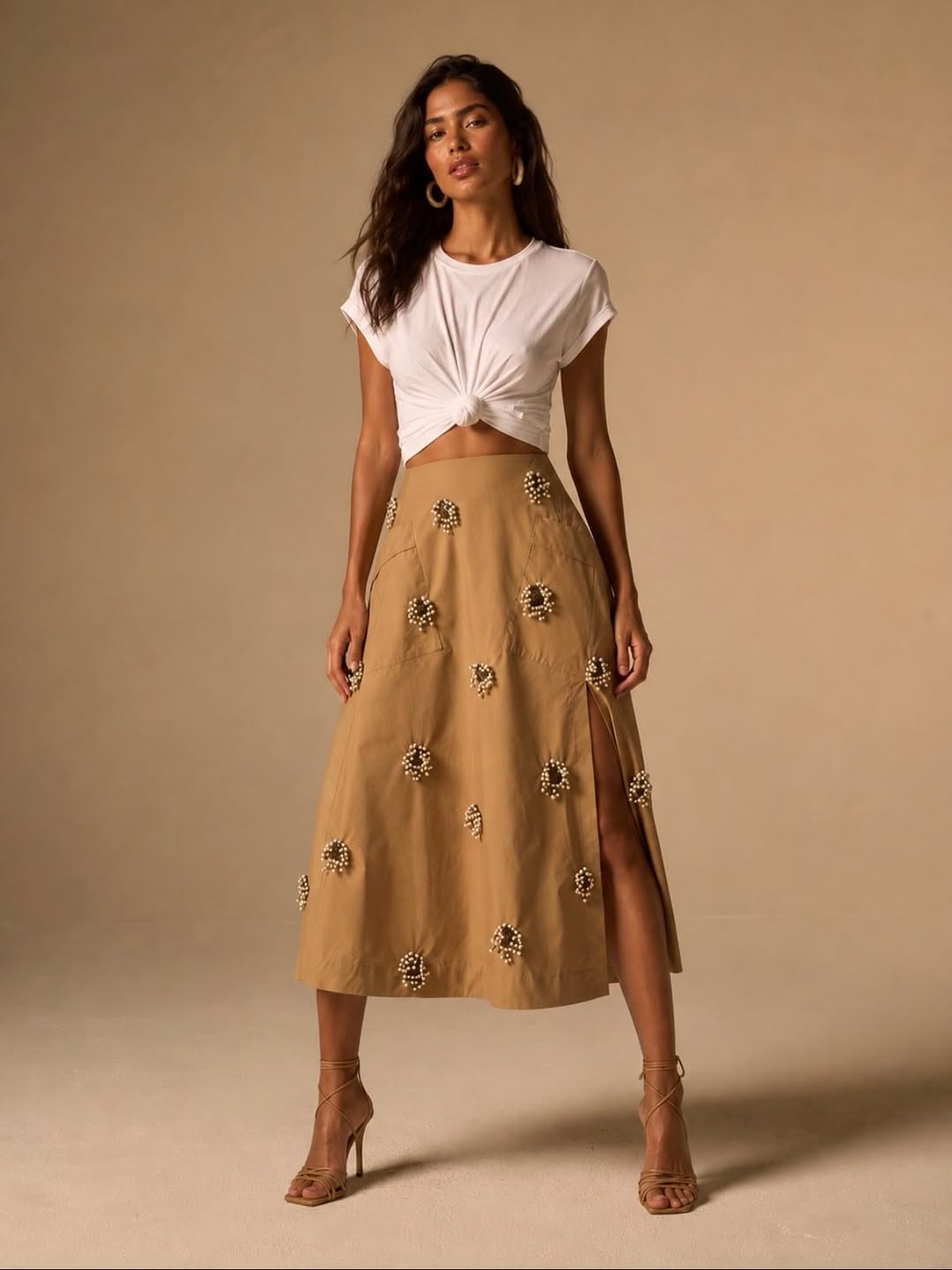 Full-body shot of a model wearing a tan A-line midi skirt with scattered pearl flower appliqués and a side slit, paired with a white knotted t-shirt.
