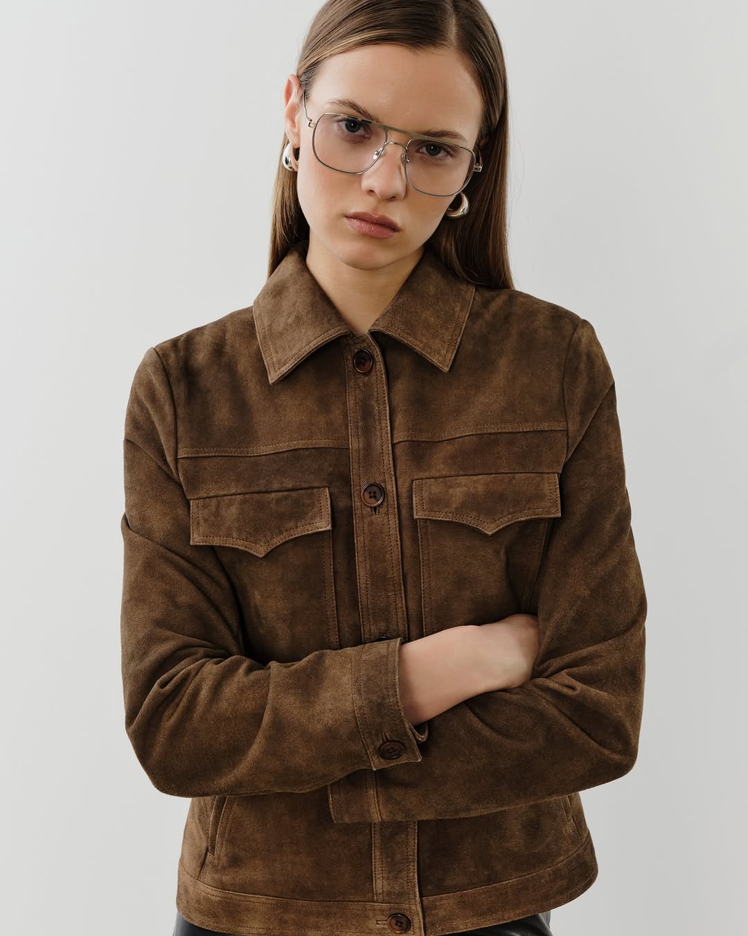 A brown suede trucker-style jacket with a pointed collar, button-front closure, two chest flap pockets, and structured seaming.