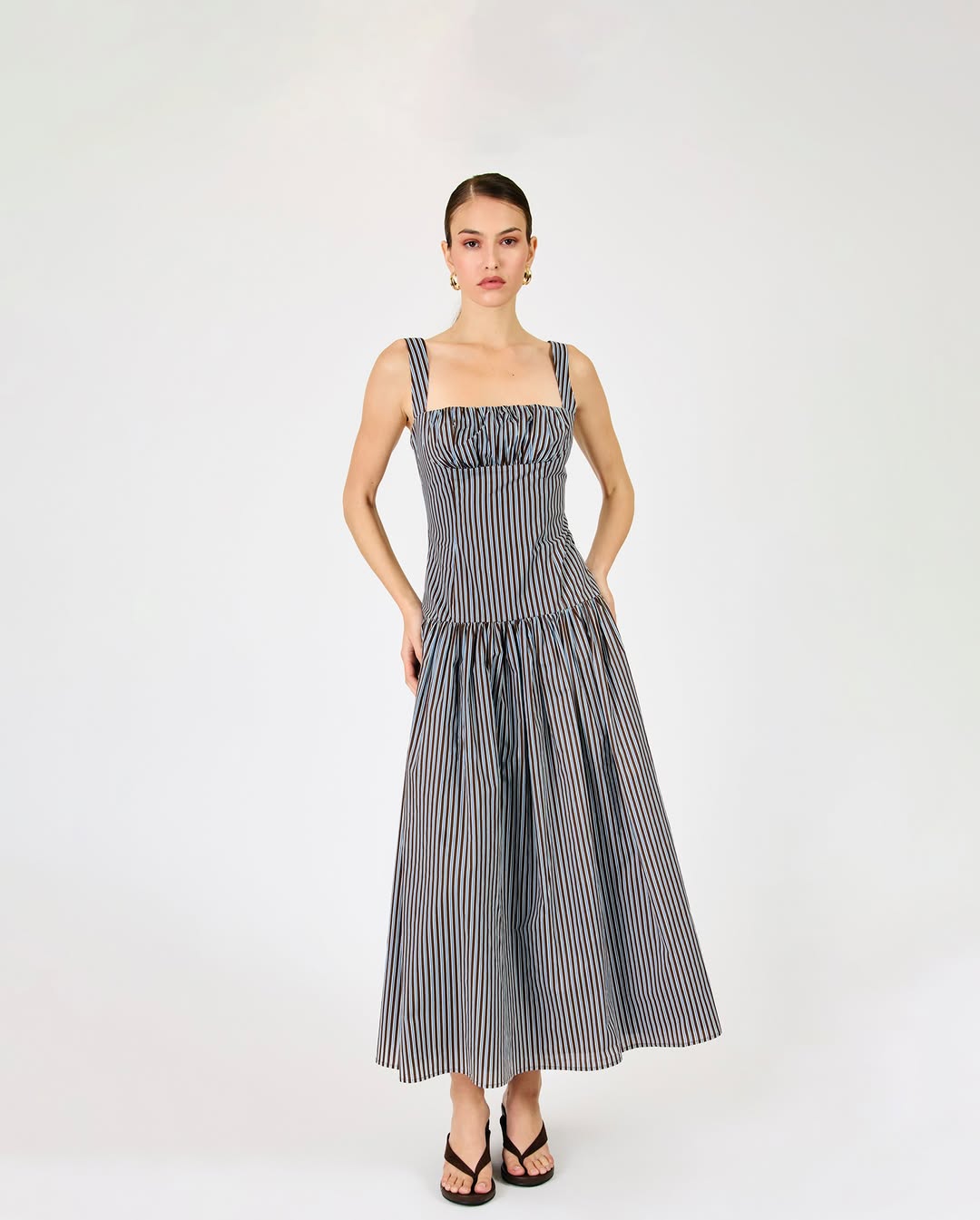 Full-body shot of a model wearing a brown and blue vertically striped maxi dress with square neckline, wide straps, smocked bodice, and tiered skirt.