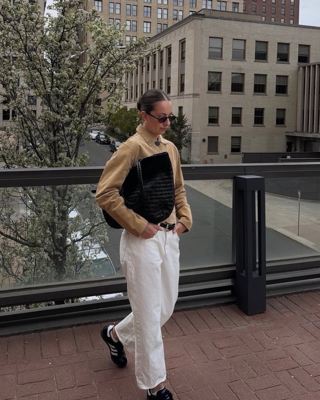 Full-body shot of a woman wearing a cropped tan suede jacket with large dark buttons over white wide-leg trousers, carrying a black woven tote bag.
