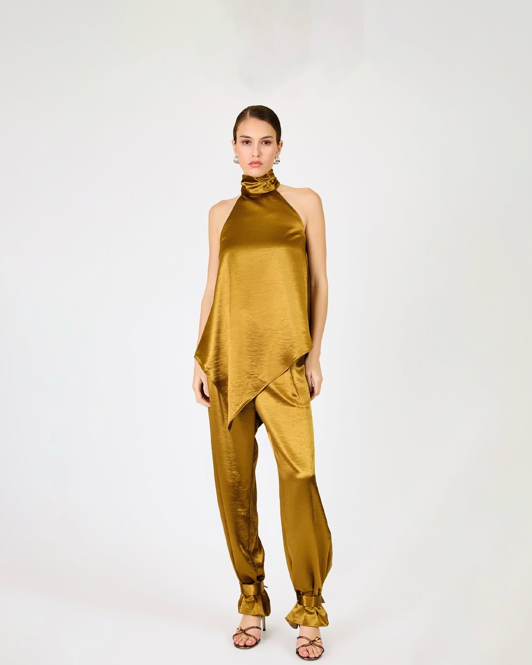 Full-body shot of a model wearing a shiny gold hal-neck top with an asymmetrical hem and matching loose trousers with ruffled cuffs.