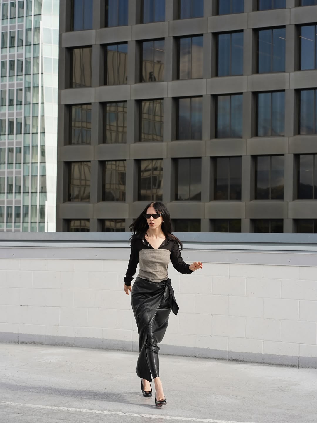 Model walking in a coordinated look featuring a black sheer long-sleeve shirt layered over a grey scoop-neck tank top, paired with a black midi-length faux leather wrap skirt…