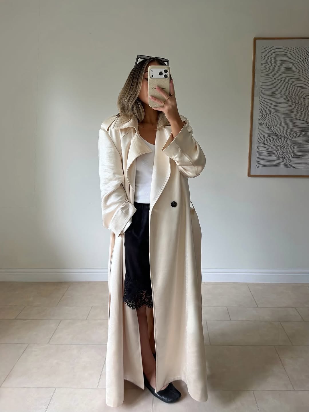 Full-length mirror selfie showing a model wearing an oversized cream satin trench coat over a white tank top and black lace-trimmed slip skirt.