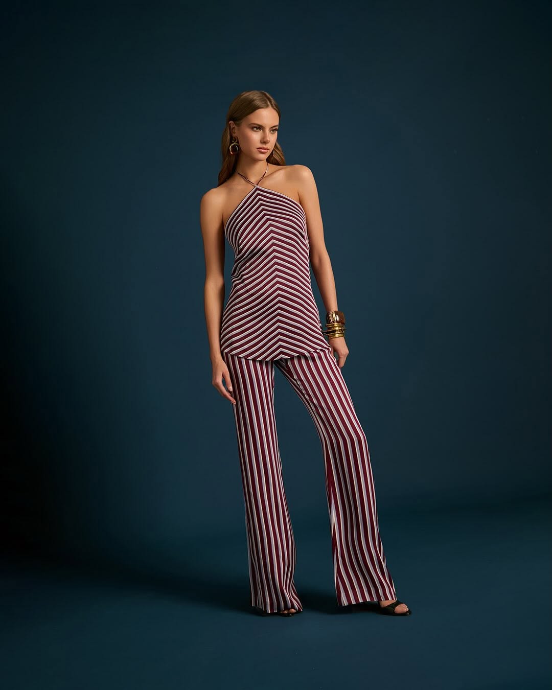 Full-body shot of a model wearing a burgundy and white striped halter top with chevron detailing on the bodice and vertical stripes on the flared trousers.