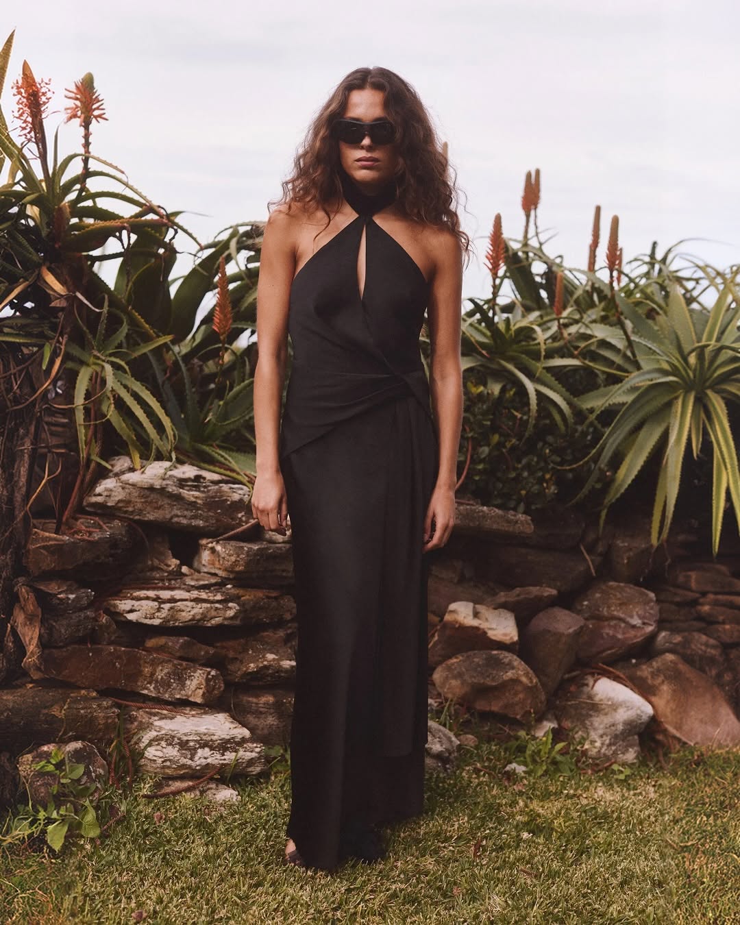 Full-length shot of a model wearing a black floor-length maxi dress featuring a high halter neckline, deep chest keyhole, and draped waist detail.