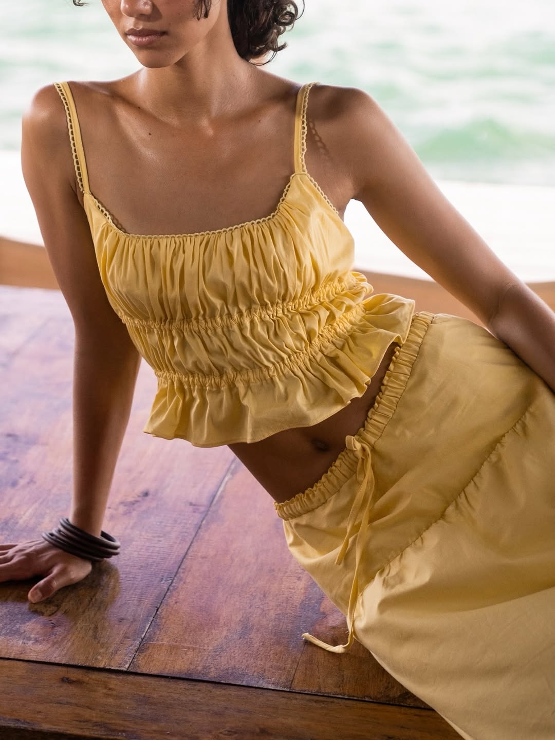 Close-up view of a model wearing a yellow spaghetti-strap crop top with shirring details and a ruffled hem, paired with a matching yellow skirt featuring an elastic drawstring…