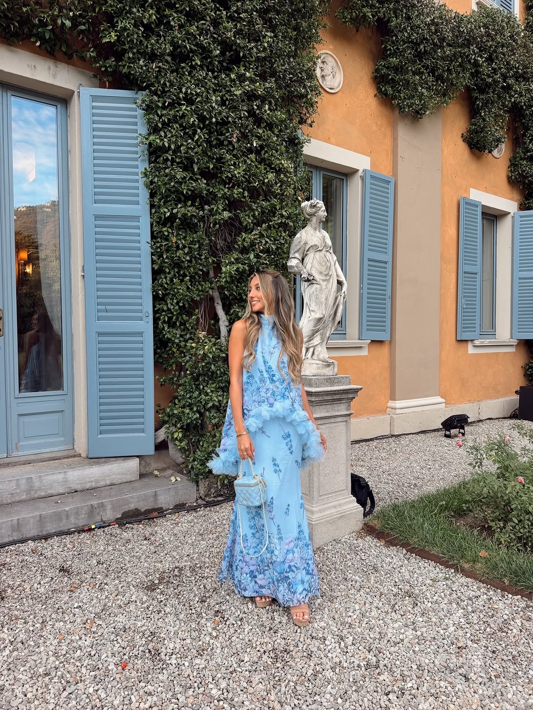 Model wearing a long, light blue halter-neck maxi dress with purple floral print and tiered tulle ruffles.
