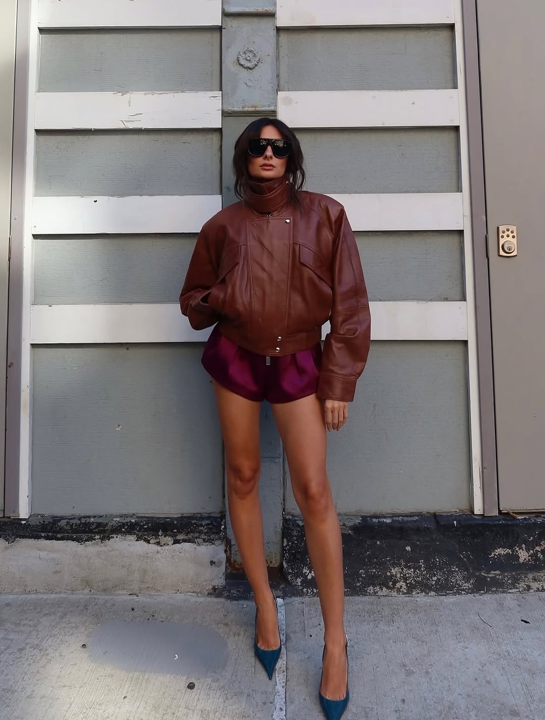 Full-body shot of a woman wearing an oversized brown leather moto jacket with a high collar, dark red satin shorts, and pointed-toe teal slingback heels.