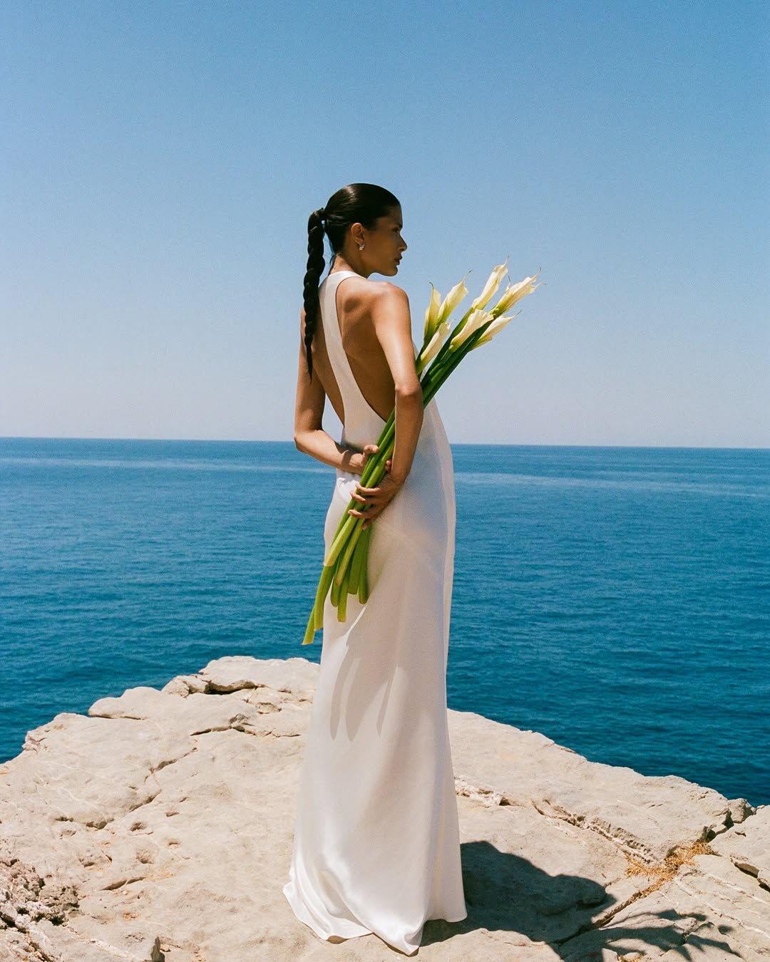 Model wearing a white floor-length gown with a low back, holding calla lilies by the sea.