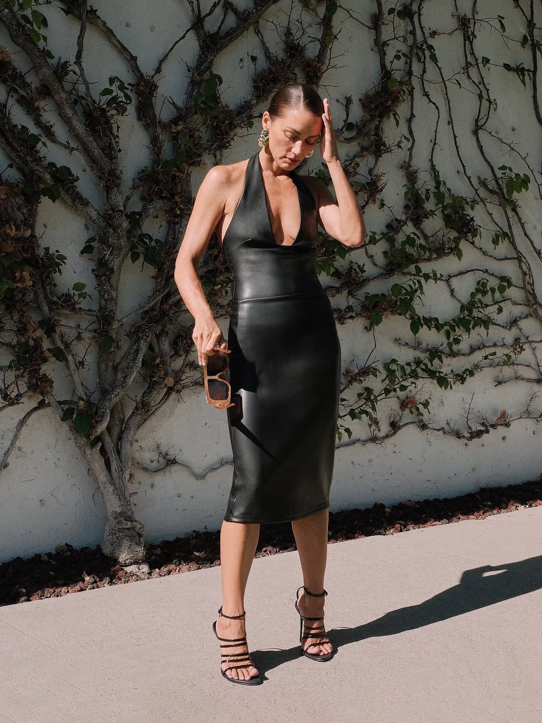 Model wearing a black deep-V halter neck faux leather midi dress with strappy heels.