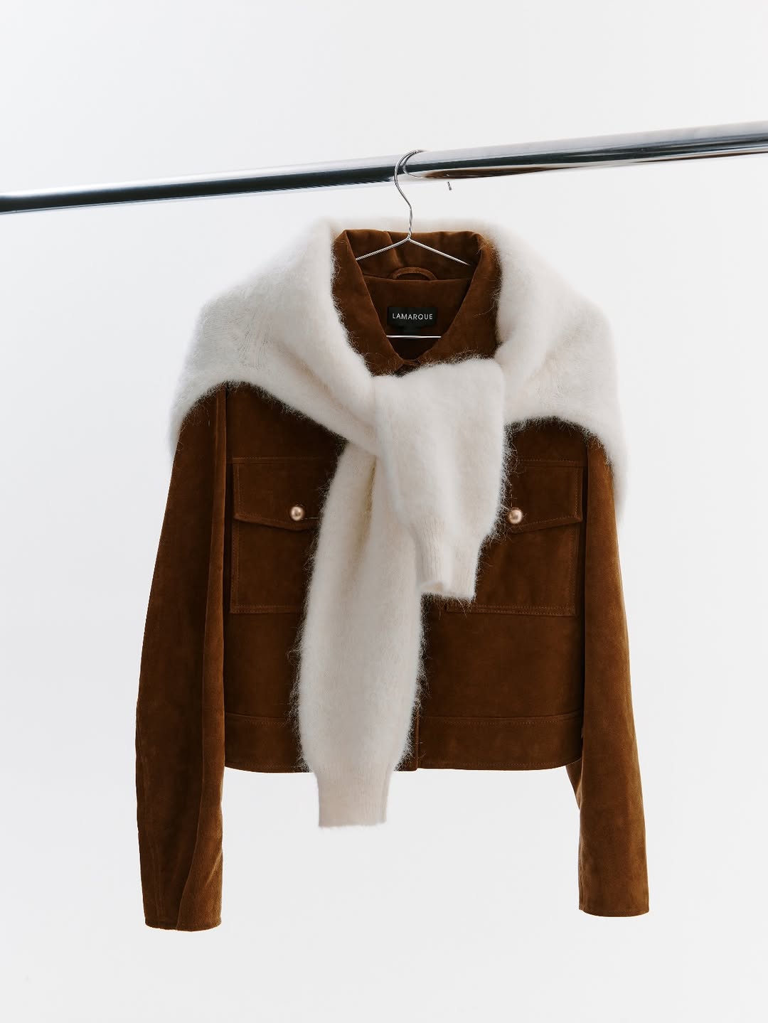 A full view of a brown suede trucker-style jacket hanging on a rack, featuring chest pockets and button details, with a cream fuzzy sweater tied around the collar.