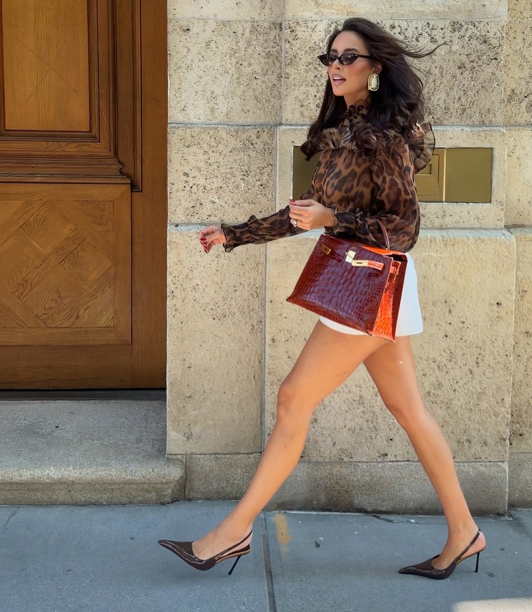 Side view of model wearing brown leopard print ruffled blouse, white shorts, red handbag, and heels.