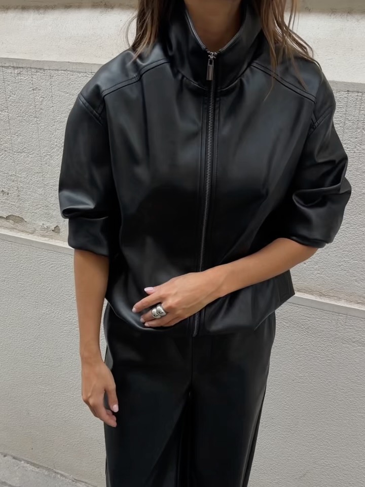 Close-up front view of a model wearing a black leather high-neck zip-up jacket with elbow-length sleeves.