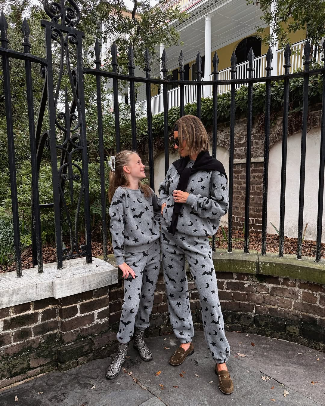 A woman and young girl stand outdoors wearing matching grey lounge sets printed with black bats, stars, and moons.