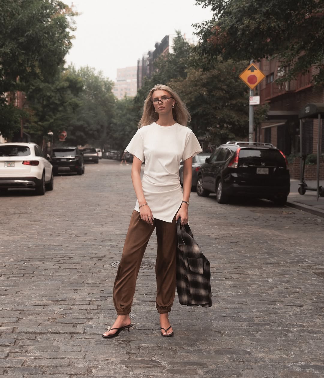 Full-body shot of a model on a cobblestone street wearing a long white t-shirt, brown joggers, and holding a plaid shirt.