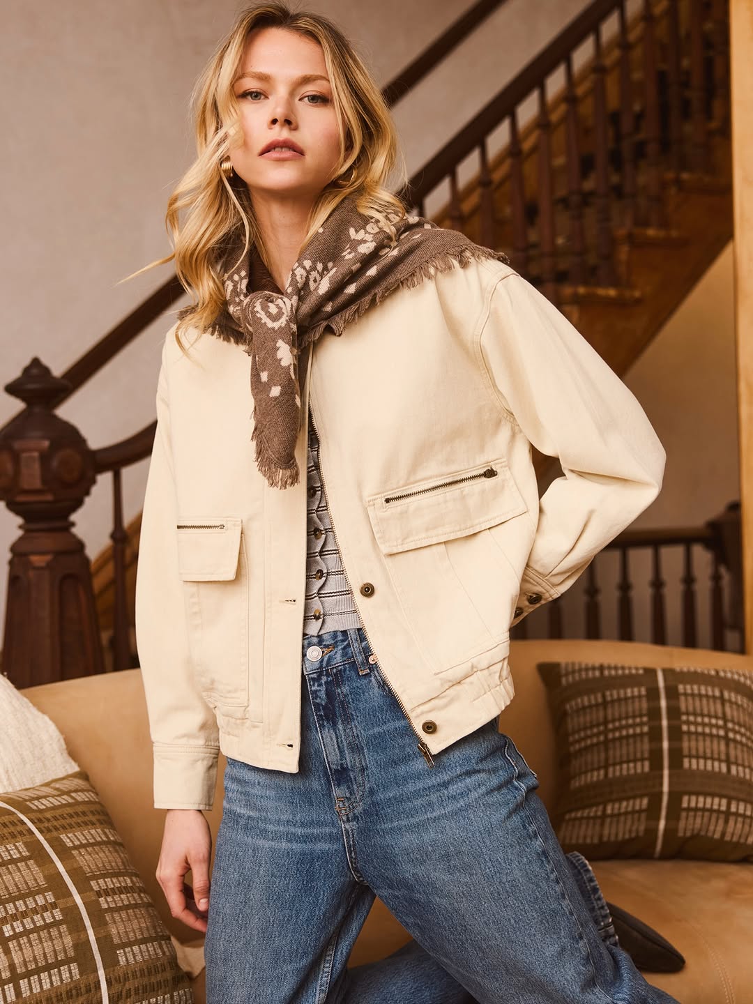 Model wearing a cream jacket and blue jeans with a brown patterned scarf draped around her neck.