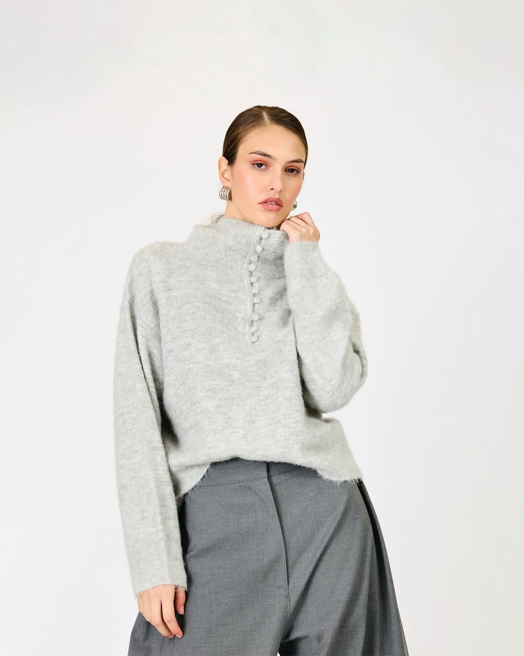 Model wearing a fuzzy light grey sweater with a row of pom-pom buttons down the front placket and wide-leg grey trousers.