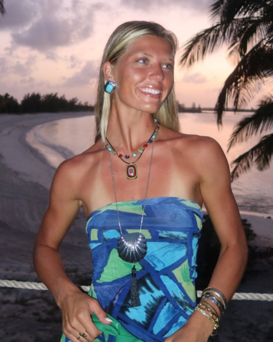 A full-body shot of a woman on a beach at dusk wearing a blue and green abstract print strapless top wrapped around her torso.